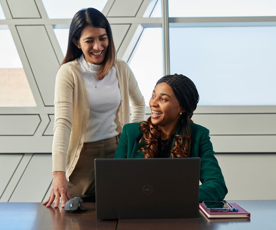 Two colleagues collaborating at a desk, with one seated using a laptop and the other standing beside them in a bright, modern office setting.