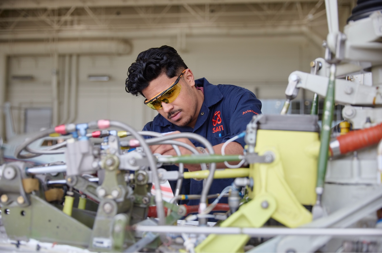 Student wearing safety glasses working on aircraft components in an aviation lab – inspecting equipment surrounded by mechanical systems and tubing.