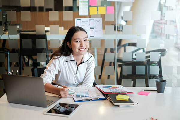 A young woman sits at a desk, focused on her open laptop.  She's wearing a casual top, with one hand on the keyboard and the other resting on the desk. The setting appears to be a bright indoor work- or study-space, with soft ambient light and minimal background distractions.