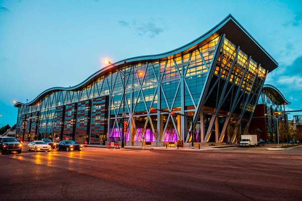 A sweeping dusk view of the modern Aldred Centre: glass-fronted with tall diagonal structural supports, interior lights glowing warmly as the sky fades to twilight. The building stands against a quiet street, its reflective surfaces and sharp roofline creating a striking architectural silhouette.