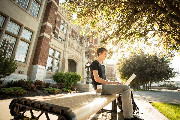 A young man sits on a bench outdoors in the warm light of late afternoon, working on his laptop. He’s facing right, the sun filtering through the leaves of a tree overhead. Behind him is a stately brick and stone building and a pathway lined with trees, suggesting a campus or institutional setting.