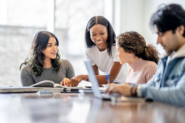 Four college-aged students sit together around a table with books and laptops. One student, standing, leans in to point something out in an open textbook while the others listen and look on. The background is softly blurred, focusing attention on the group’s engaged collaboration.