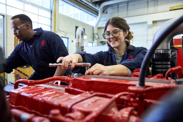 Two people wearing safety glasses work on a large red engine in a workshop.