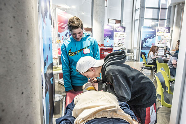 Several youth participants kneel on the floor around a training manikin in a classroom setting, one student pressing both hands on the manikin’s chest to simulate chest compressions while others watch and prepare to follow — conveying an engaged group CPR practice session.