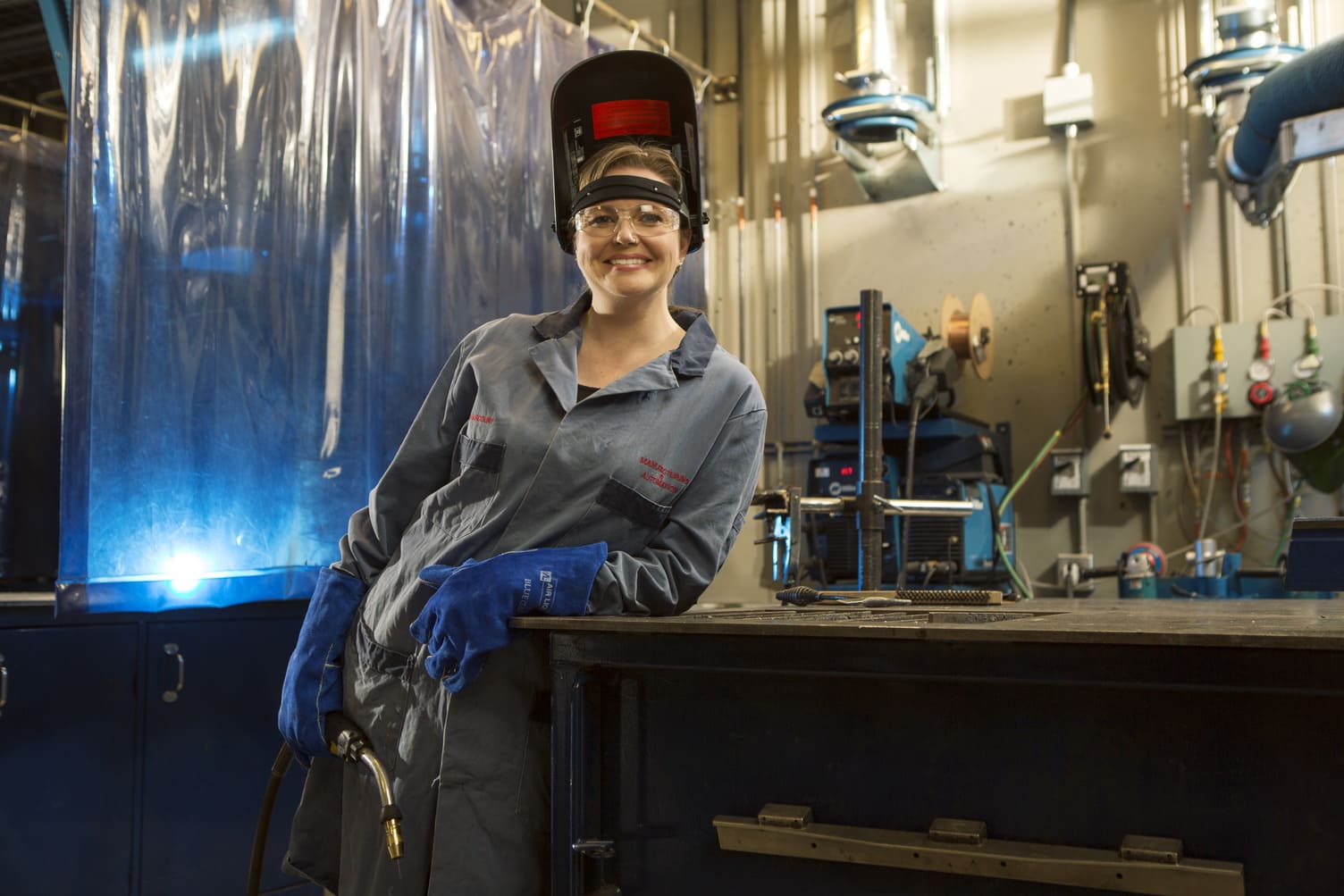 A woman wearing coveralls and a welding helmet poses for a photograph in a SAIT welding lab.