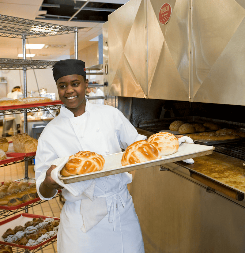 A smiling baker holds a tray of freshly baked bread in a commercial kitchen. The oven is visible in the background, showcasing the baking process.