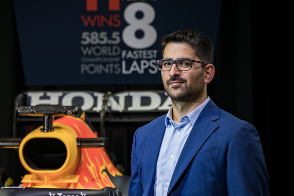 Man in a blue suit and glasses standing in front of a Red Bull Formula 1 car and Honda signage.