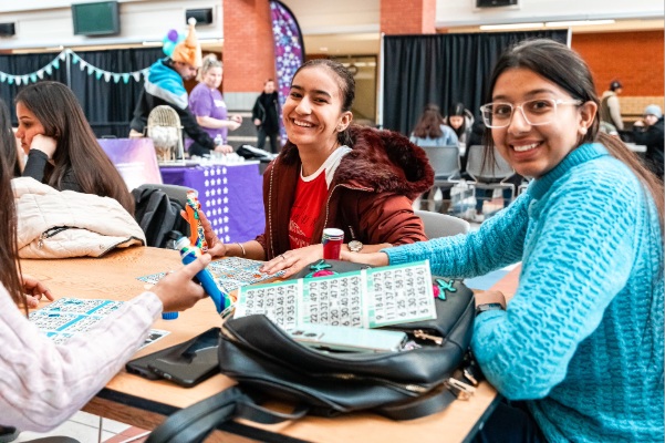 Three students sitting at a table at a campus event, smiling while playing bingo, with other activities and booths visible in the background.