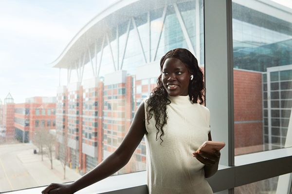 Person standing by a large window holding a phone, with a modern campus-style building visible outside.