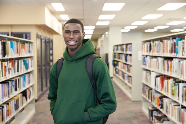 A student smiles directly at the camera while standing in a library, with shelves of books and soft overhead lighting behind them—conveying a warm and inviting academic environment.