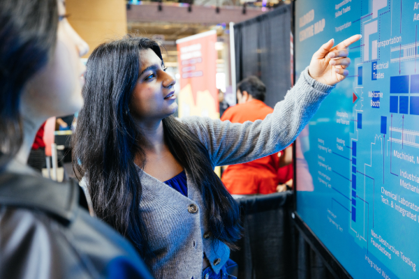 Person viewing information on a large display screen at an event booth – pointing to a campus map while discussing program areas with another attendee.