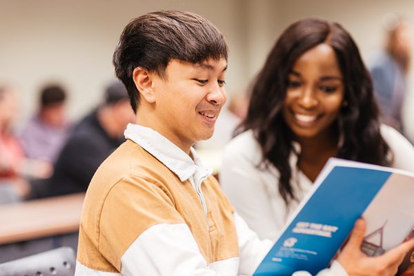Two people sit side by side in a classroom setting, with one holding and reading a booklet.