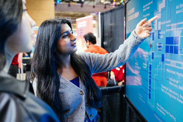 Two people stand at a booth while one points to a large digital display showing a campus.