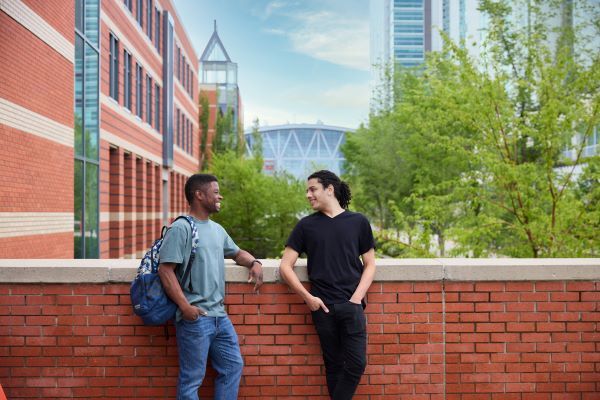 Two students have a conversation in front of a brick wall on campus, smiling.