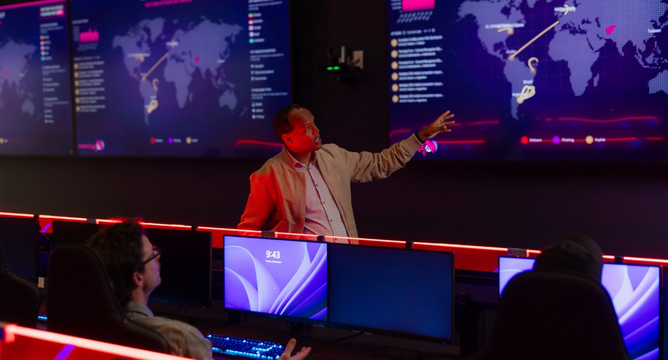 A man gestures towards a digital world map displayed on multiple screens while explaining data to two seated individuals in a high-tech environment.