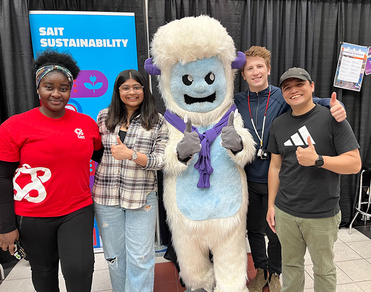 Photograph shows four people posing with a smiling white-and-blue mascot at a SAIT sustainability event. Blue sustainability banner, purple accents, thumbs-up gestures, and event materials frame group.