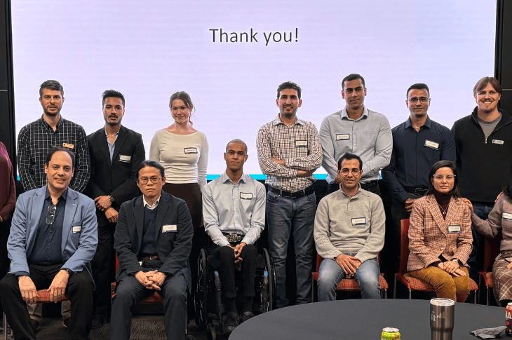 A group of people, some sitting, some standing, gather in front of a screen that reads "Thank you!" in black text on a white background. They are all looking at the camera and smiling. 