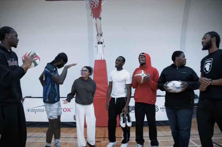 A group of eight individuals stand beneath a basketball hoop in a gym. Some hold sports balls including a volleyball, basketball, and rugby ball. They appear to be talking casually and interacting with one another.