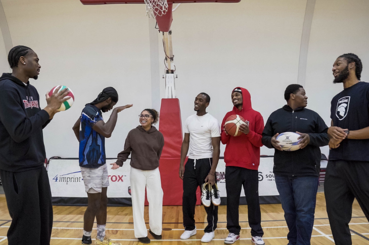 A group of eight individuals stand beneath a basketball hoop in a gym. Some hold sports balls including a volleyball, basketball, and rugby ball. They appear to be talking casually and interacting with one another.