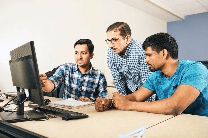 Three people gather around a computer monitor and look at something on screen.
