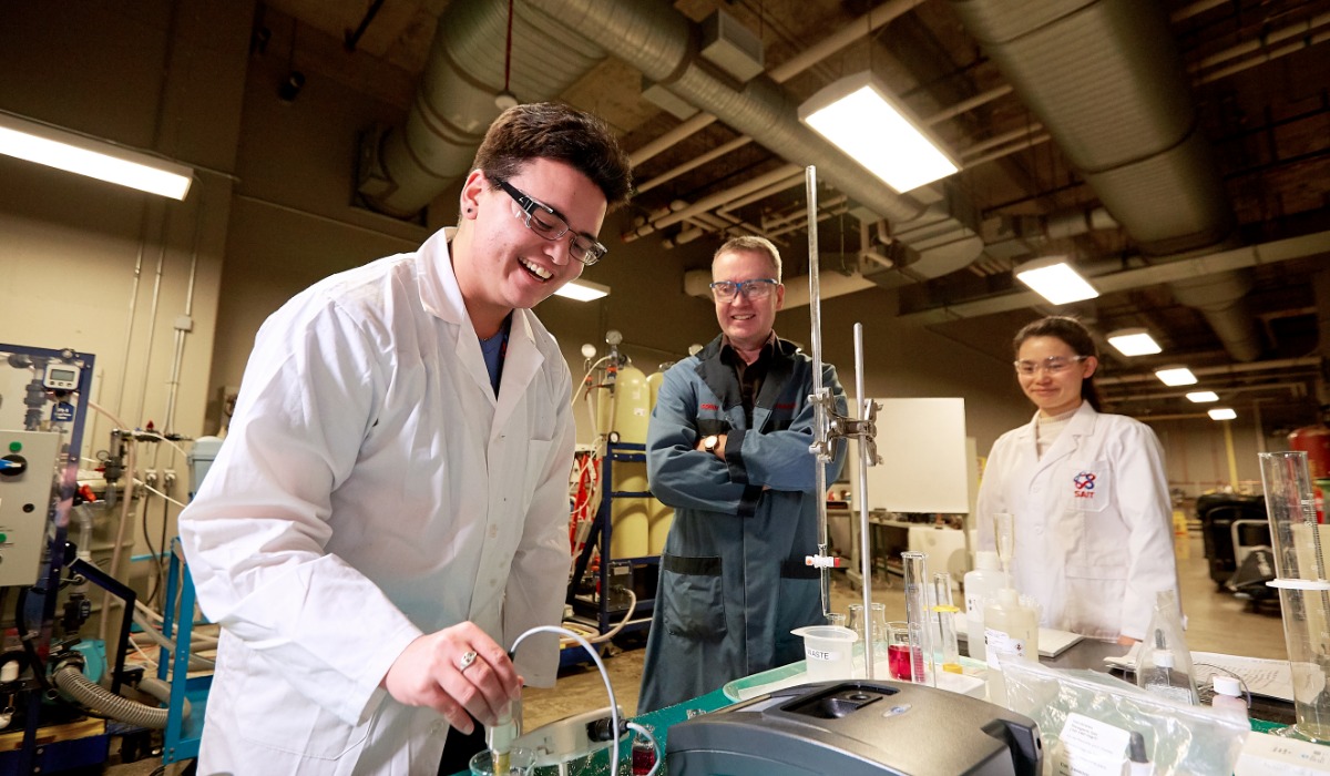 Researchers in lab coats and safety glasses conduct an experiment at a laboratory bench with measuring equipment.
