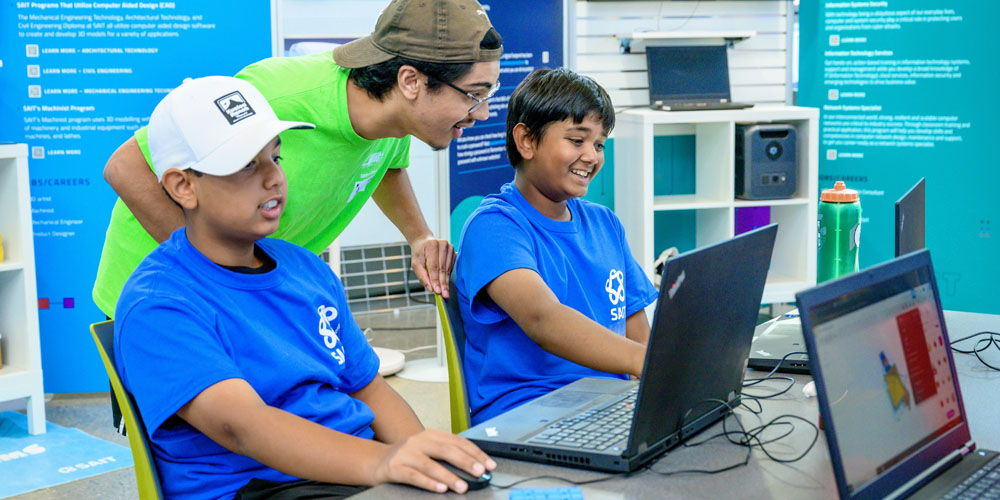 Two young boys in blue shirts are engaged with laptops while a mentor in a green shirt assists them, showcasing a collaborative learning environment.